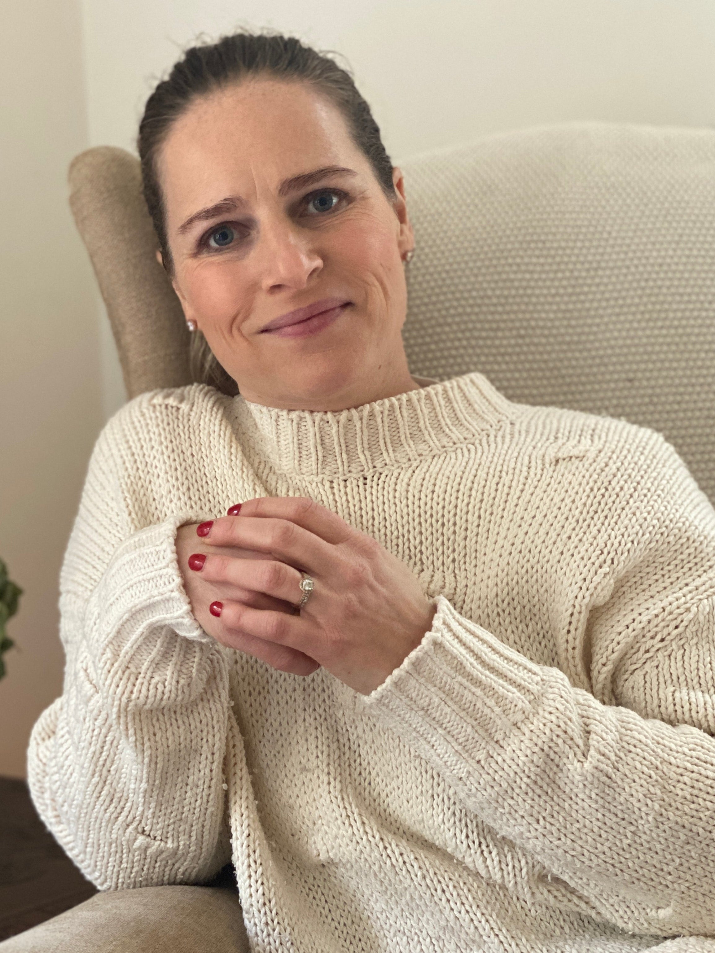 Woman sitting on a couch wearing a white sweater, holding her hands together
